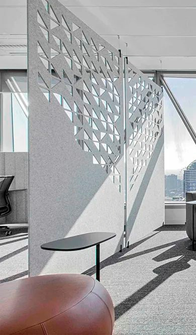 Open plan office with geometric partition screens creating private work nooks natural light streaming through windows and a round orange ottoman with a small side table