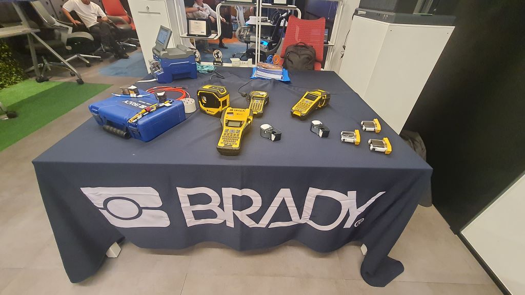 Table with a BRADY banner displays yellow handheld labeling/identification devices and a blue case on a dark tablecloth in an exhibition setting.