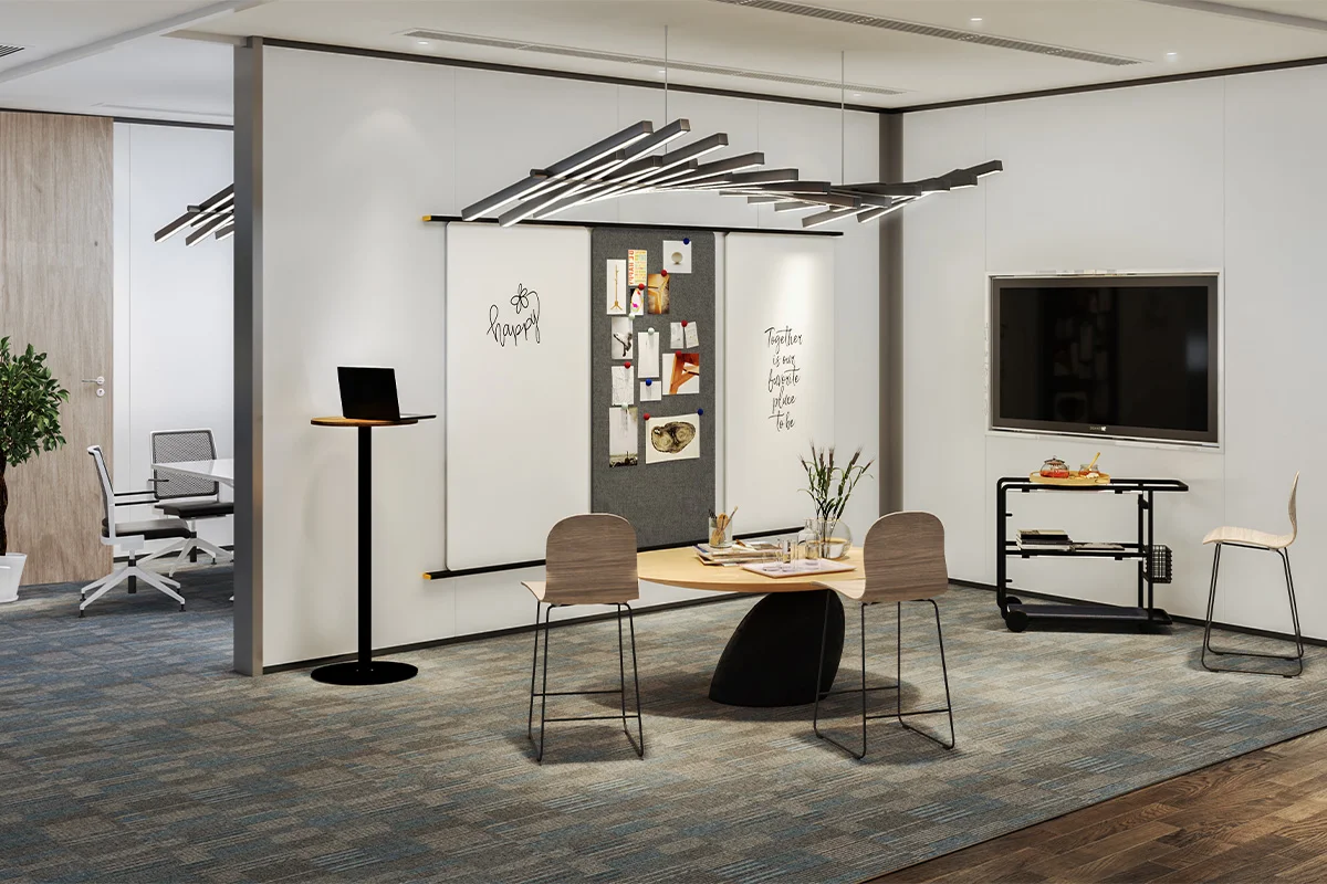 Open-plan office breakout area with a round wooden table and three chairs on a patterned carpet, opposite a whiteboard wall and a wall-mounted TV on the right.