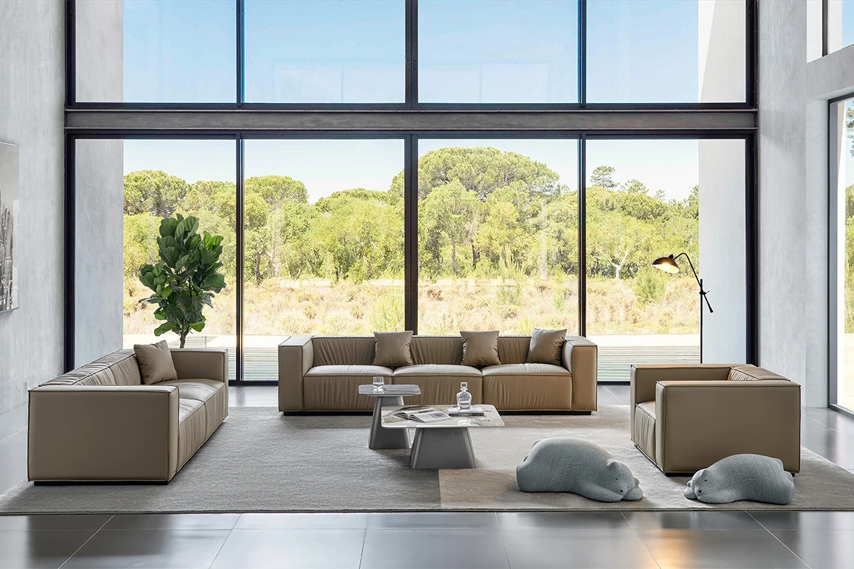 Modern living room with large floor-to-ceiling windows showing a green landscape; beige leather sofas, a gray area rug, plants, and a floor lamp. Two plush animal cushions rest on the rug.