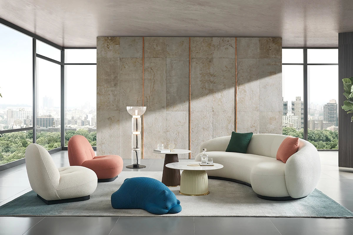 Modern living room with a curved white sofa, pink and green cushions, blue pouf, and round coffee tables; floor-to-ceiling windows show a city skyline.