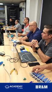 Five men seated at a long table in a tech workshop, with laptops, cables, and network gear in use; a server rack in the background and a branded banner at the bottom.