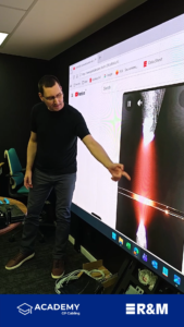 Instructor in a black shirt points at a large display during a tech training session, with a browser window visible on the screen.