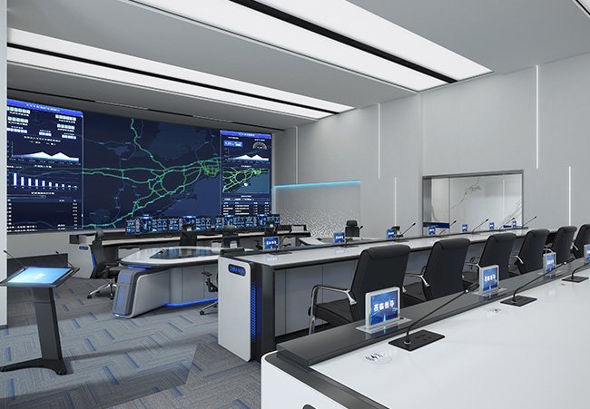 Control room with large wall screens showing maps and charts, and rows of desks with computer monitors and chairs.
