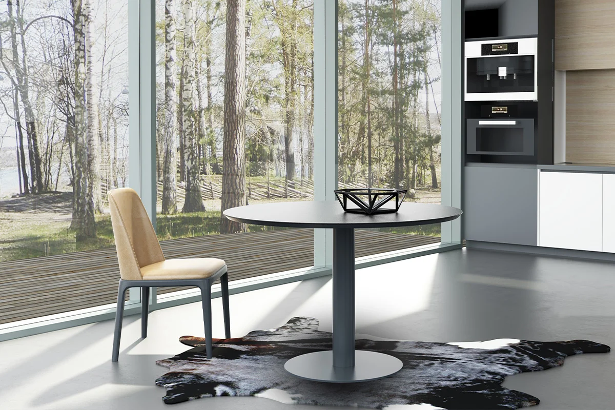 Modern dining area with a round gray table, beige upholstered chair, and a wall of glass windows overlooking a wooded outdoor scene.