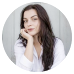 Portrait of a woman with long dark hair in a white shirt resting her chin on her hand and looking softly at the camera