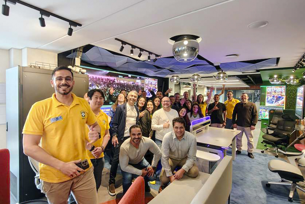 Diverse group of coworkers posing for a group photo in a modern, colorful office or studio space, with several people smiling and giving thumbs up in the foreground.