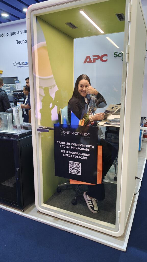Smiling woman inside a glass booth at a tech expo giving a thumbs up with a One Stop Shop sign and QR code poster nearby