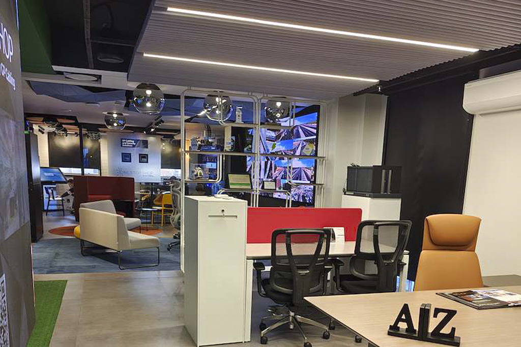 Open-plan modern office with a reception desk, glass partition, and lounge seating; orange chair and a desk with AIZ letters visible in the foreground.