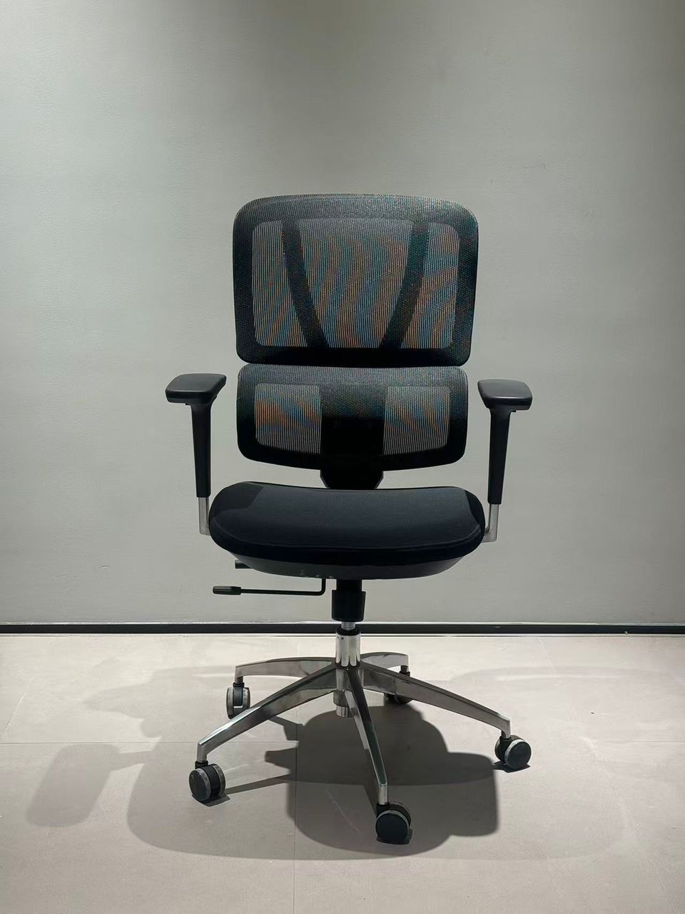Ergonomic office chair with a black mesh back, cushioned seat, adjustable armrests, and a five-star wheeled base against a gray wall.