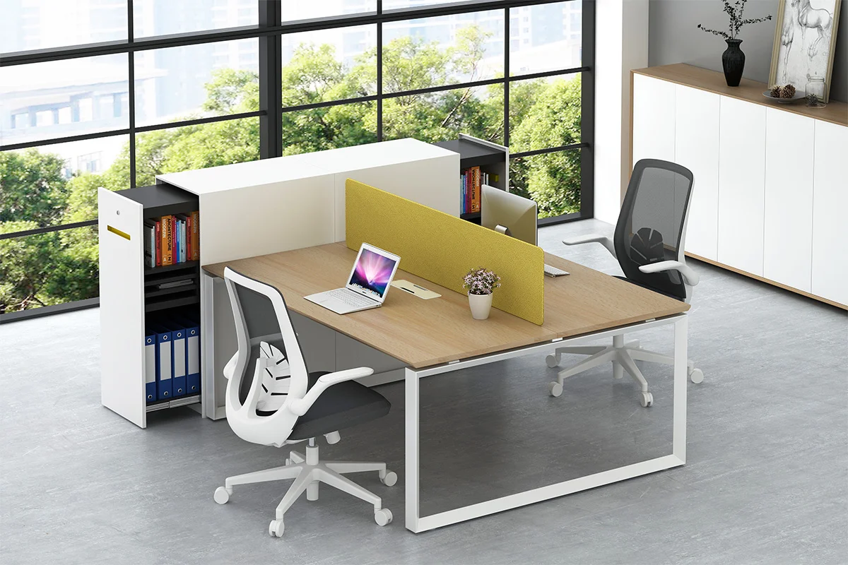 Open plan office with two desks arranged side by side separated by a yellow divider facing large windows with greenery outside A laptop and plant sit on the desk