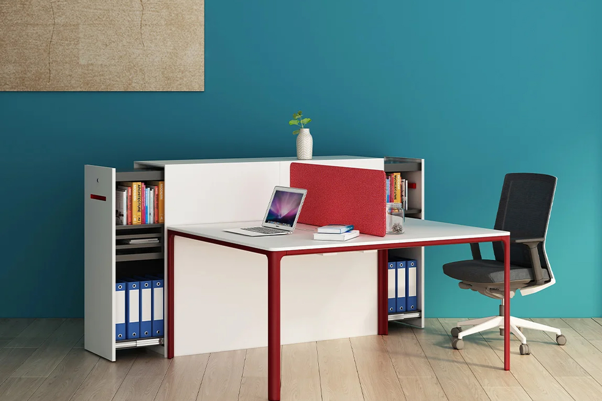 Modern office workspace with a white desk, red legs and divider, laptop on the surface, a small plant on top, and shelves with books and blue binders against a teal wall.