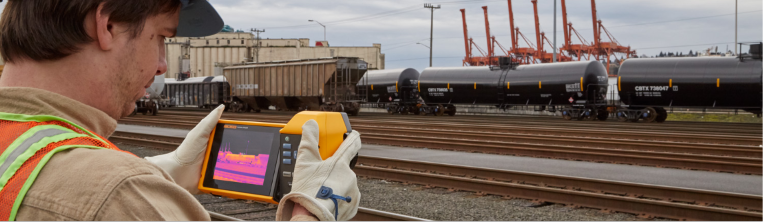 Rail yard worker in safety vest using handheld infrared camera to inspect tank cars on tracks