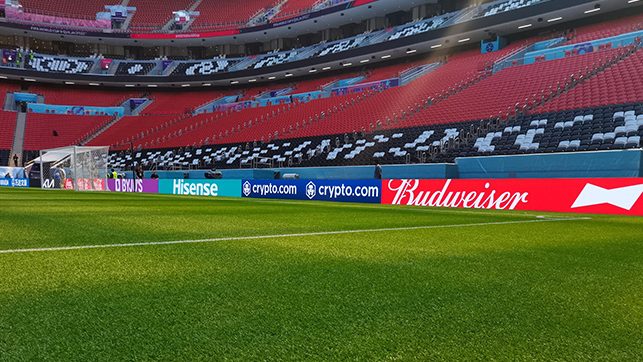 Empty stadium with red seats and a green soccer pitch; advertising boards along the sideline display Budweiser and Hisense logos.