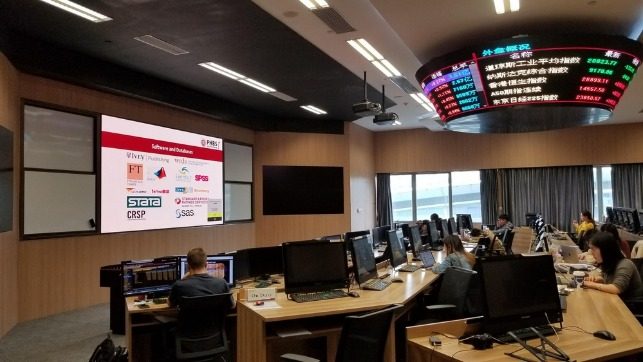 Trading room with multiple workstations and monitors, a large presentation screen showing logos, and an overhead stock ticker board.