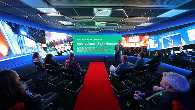 Speaker presenting to an audience in a dark auditorium, with a large screen showing 'Audiovisual Experience' and a red carpet leading to the stage.