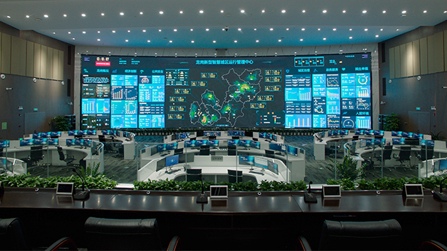 Command center with a curved desk, dozens of workstations, and a wall of blue screens displaying maps, charts, and dashboards.
