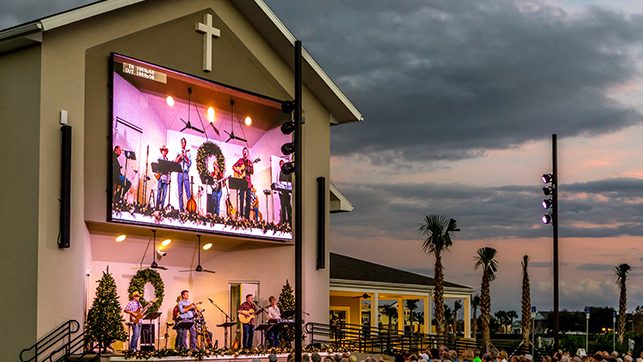 Outdoor church venue at sunset with a large LED screen showing a live band performing on stage; wreaths and Christmas lights decorate the area.