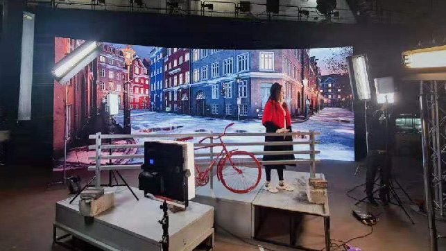 Film studio with a cityscape backdrop; a woman in a red jacket stands beside a red bicycle on a raised platform, surrounded by lighting and filming gear.