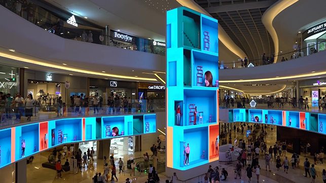 Busy multi-level shopping mall with a tall blue illuminated display tower and blue-lit storefronts, shoppers in the atrium.