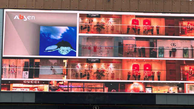 Wide digital billboard showing a multi-store fashion mall facade with illuminated shop windows and sale signs, including recognizable brand logos like Gucci and Armani.