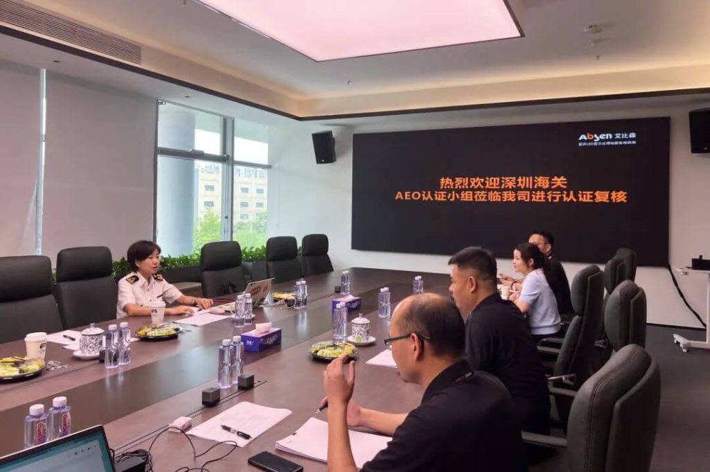 Conference room scene with people seated around a long table; a woman in a uniform speaks at the head of the table, while a screen with Chinese text is on the wall behind.