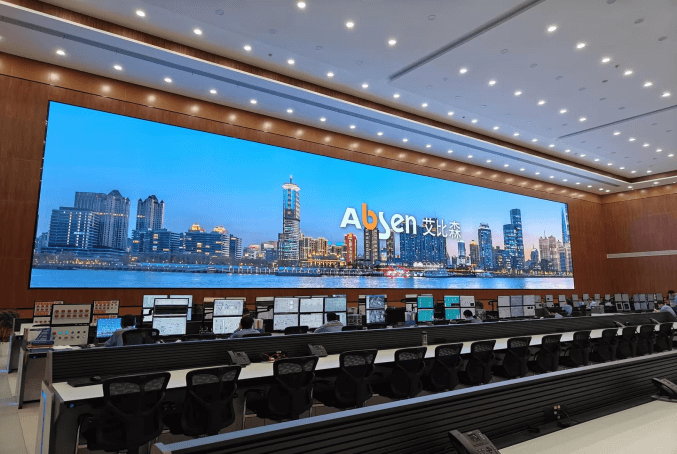 Large control room with a bright, wall-sized screen showing a city skyline and the 'Absen' logo; row of workstations and people monitor displays in front.