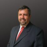 Professional head and shoulders portrait of a middle aged man in a dark suit and red tie smiling with a dark gray gradient background