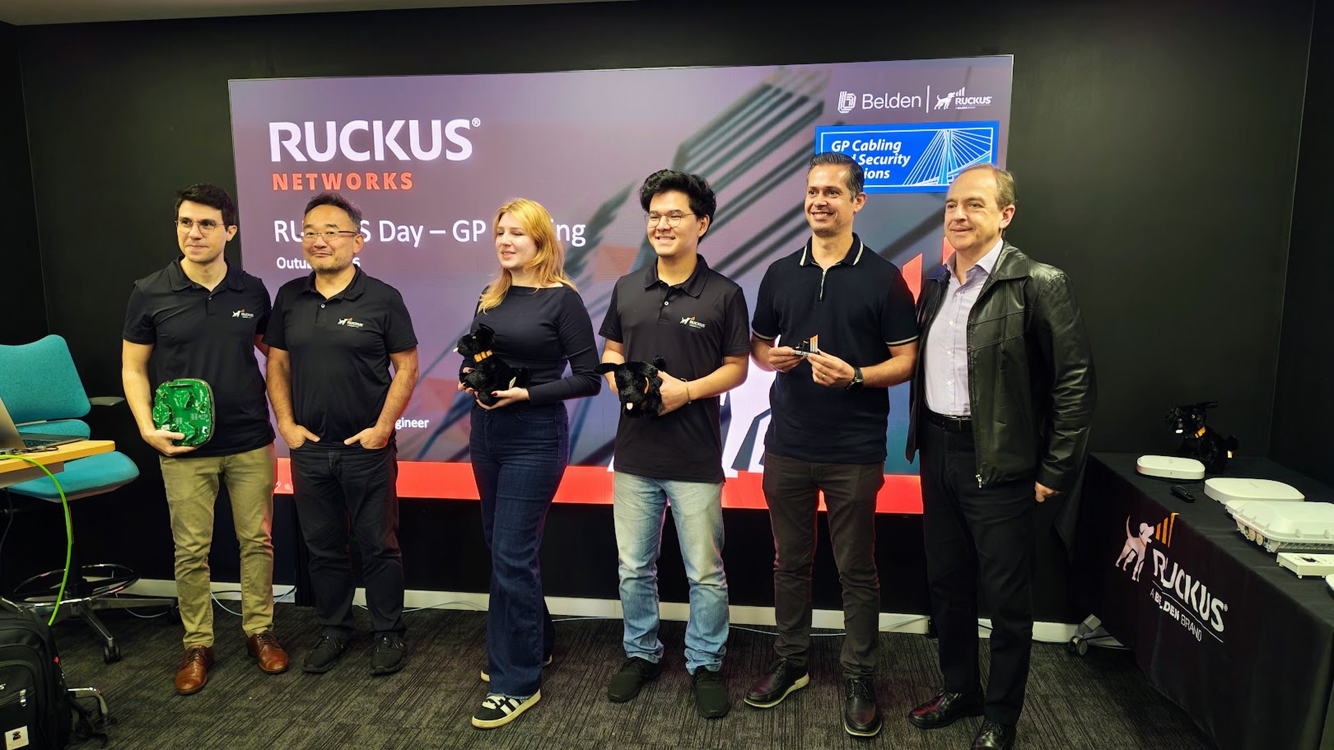 Six people pose for a photo in a tech event room, standing in front of a RUCKUS Networks banner with a blue display. They wear dark polo shirts and hold tech hardware and a plush toy.