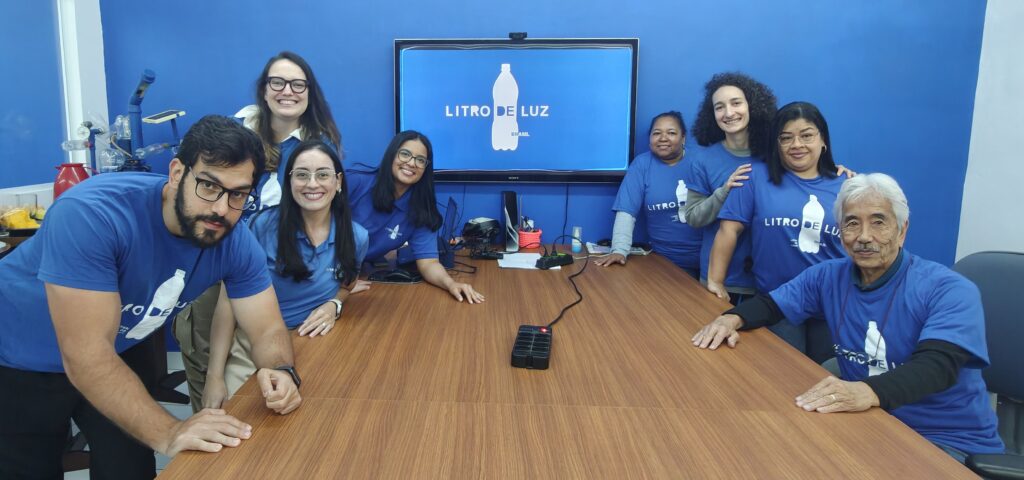 Group of diverse volunteers in blue shirts gathered around a wooden conference table smiling at the camera in a Litro de Luz Brasil room with a blue wall and screen behind them