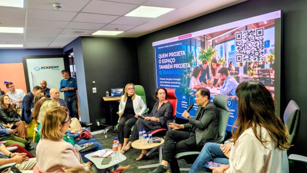 Panel discussion in a modern conference room with five speakers seated in front of a large banner audience watching from chairs and couches